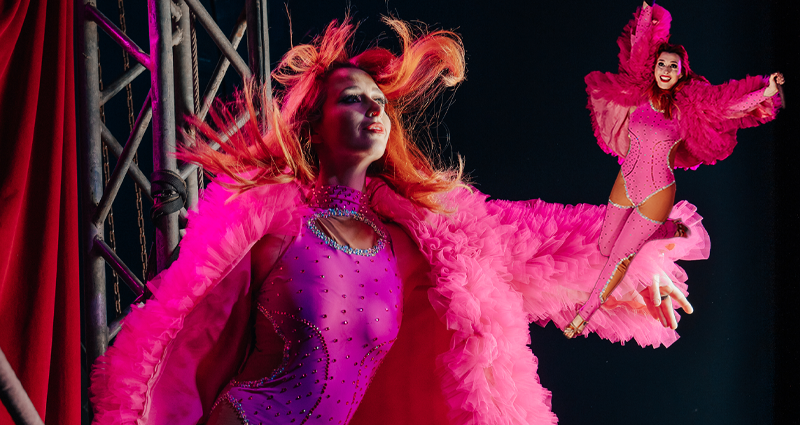 A woman in a pink leotard and pink feathery coat holds her arm out, with another woman in the smae outfit motions at the side