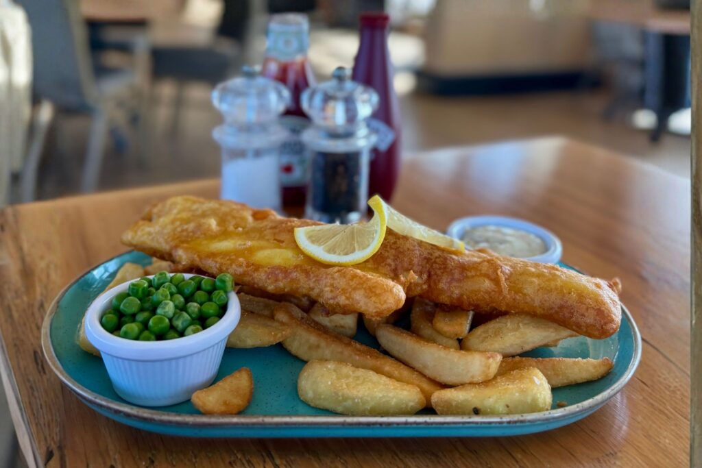 A plate of fish and chips with a slice of lemon on the fish and a pot of peas in the foreground