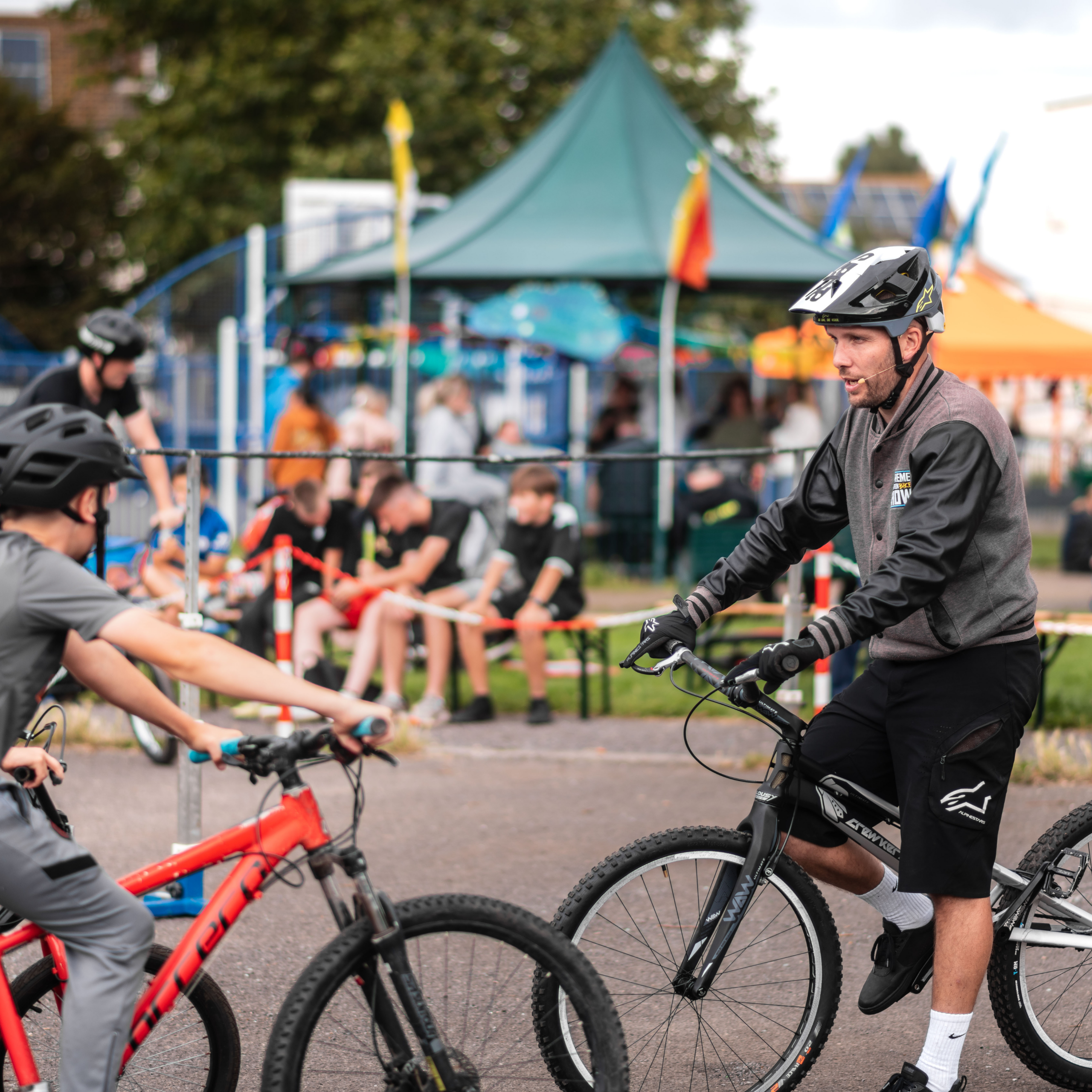 A man in biking gear and helmet sits a bike. in front of him a boy sits on a bike. Behind are various stalls and gazebos