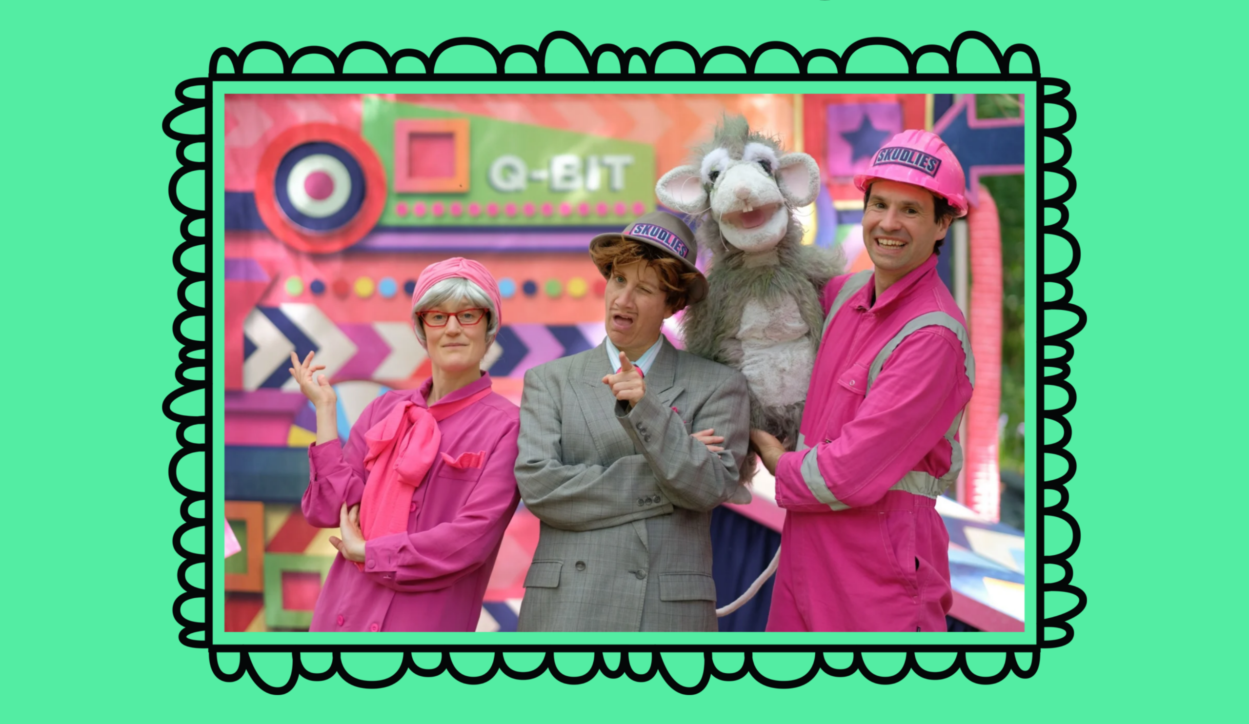 Two people wear pink safety outfits and hard hats. One holds a puppet of a giant rat. In the middle someone stands in a grey suit
