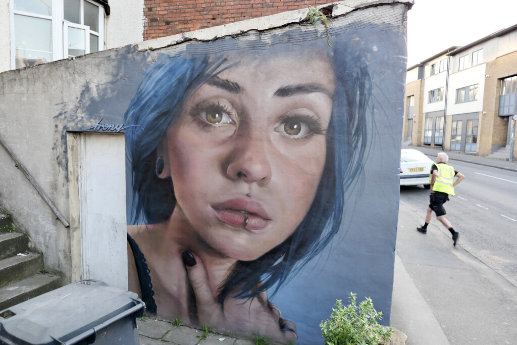 Street art portrait of the face of a lady with blue hair and face piercings painted on a wall