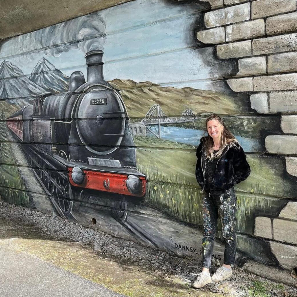 Female street artist Danksy standing in front of her mural of a black steam train cutting through mountain scenery and green fields with a river and railway bridge in the background