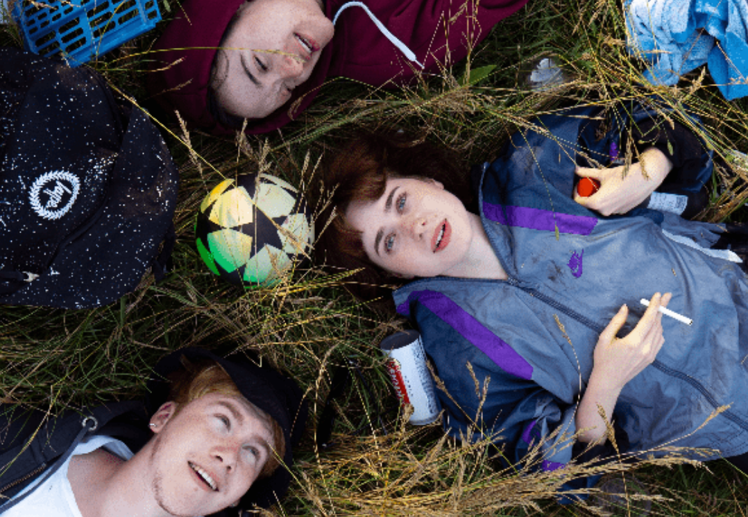 Birds eye view shot of 3 young people laying down in the grass, surrounded by clothes, a football and cigarettes