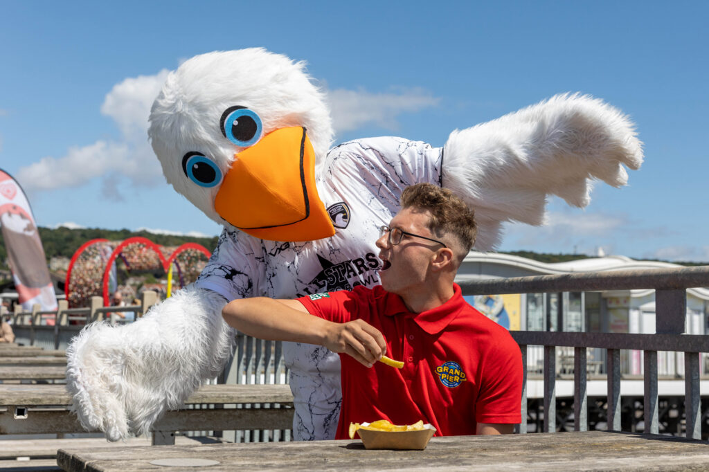 A giant seagull mascot leans over a man in a red t-shirt and glasses and tries to steal chips from the shocked-looking man