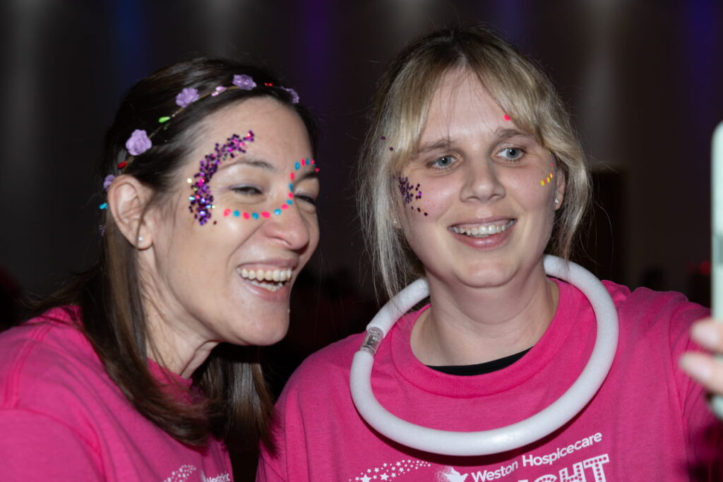 Two women wearing pink shirts and with purple glitter on their faces stand together