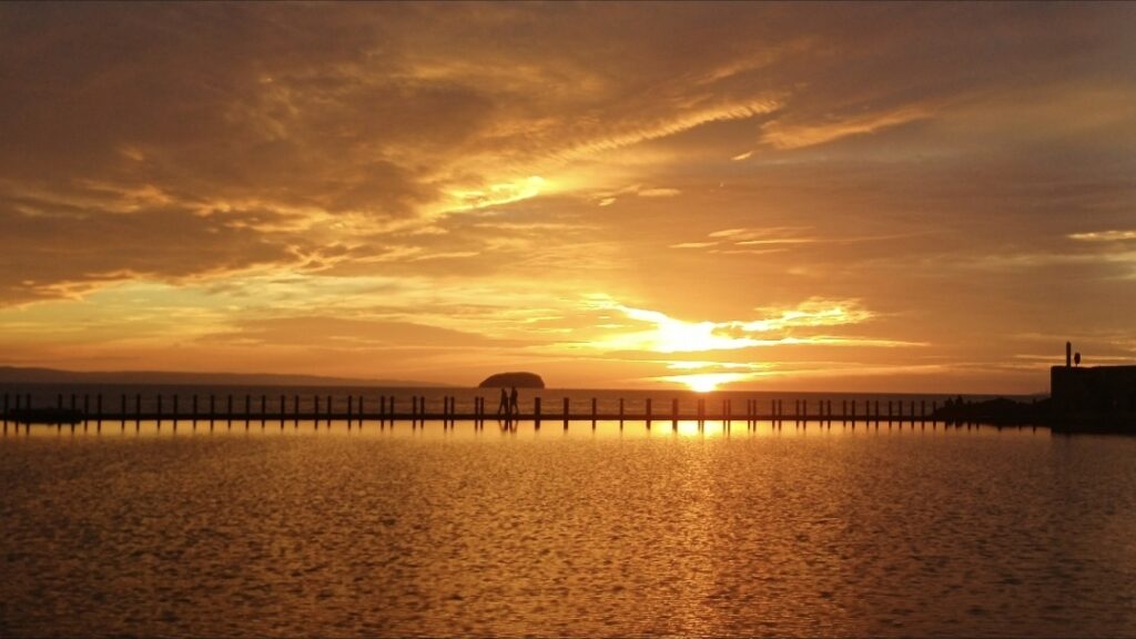 A golden sunset over a marine lake which has a fence pole lined causeway and a small island in the distance. Two silhouettes are on the causeway in front of the island