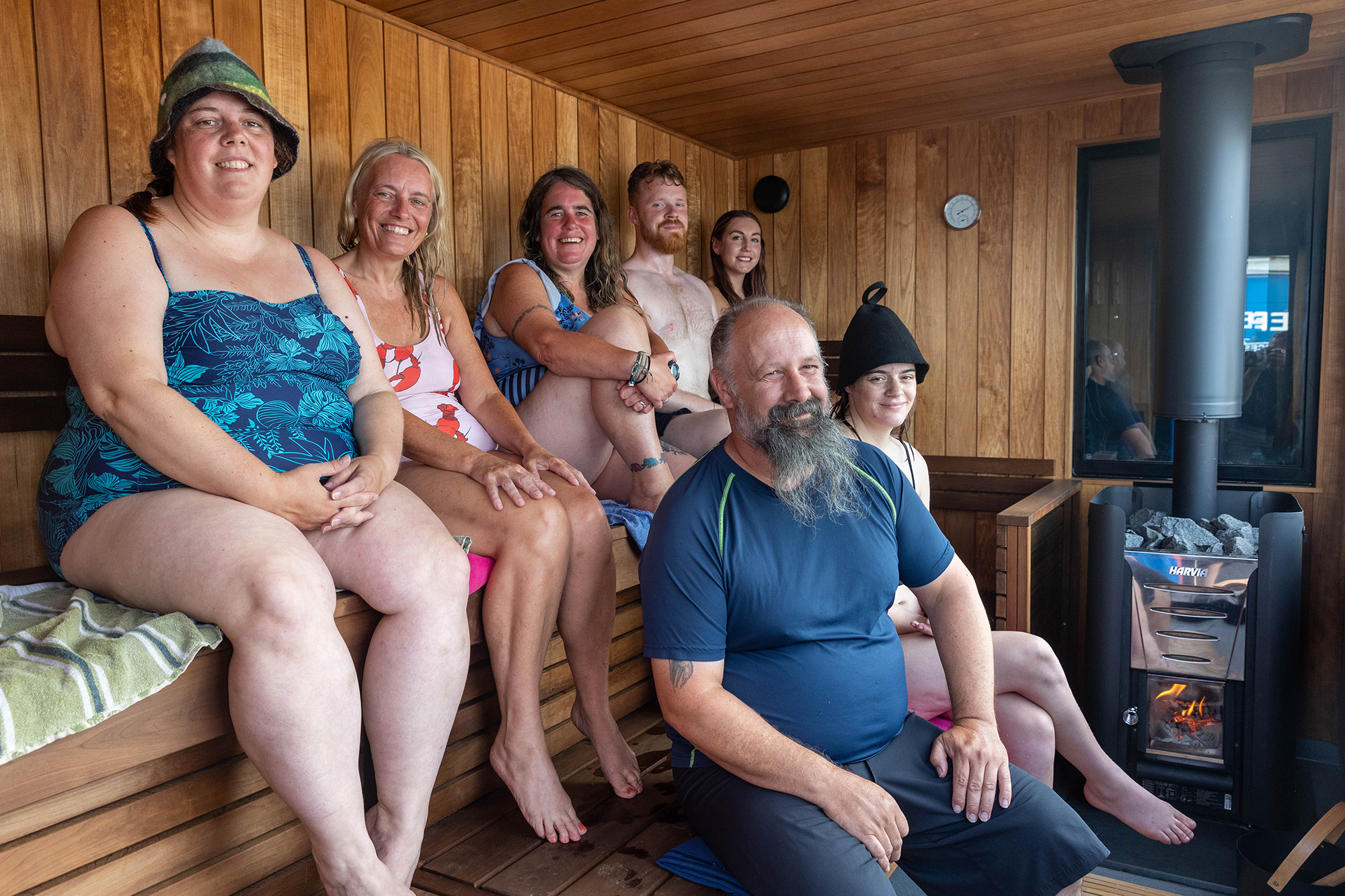 Five women and two men in swimming clothes sitting in two tiered rows inside a wooden sauna next to a flaming wood stove