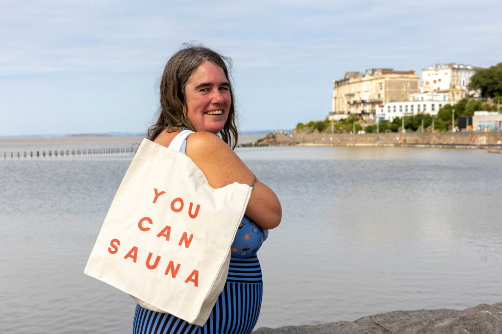 A lady in a blue swimming costume with a Tote bag on her shoulder saying You Can Sauna walks past a large marine lake