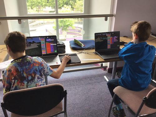 Two children sit at desks with laptops in front of them
