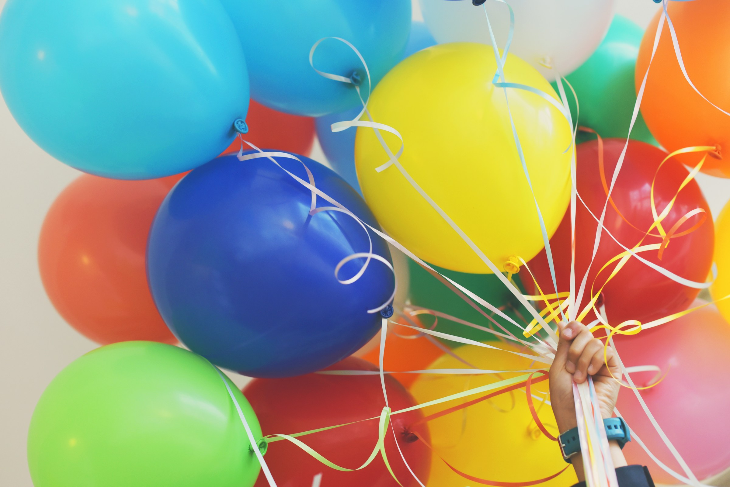 A collection of helium balloons in all different colours. They all have a white string attached to them which are held by a hand in the bottom corner