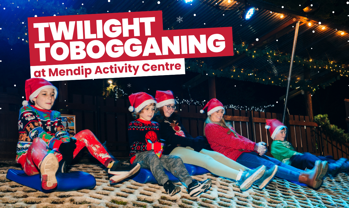A group of children sit on toboggans at the top of a slope, all in christmas hats and jumpers
