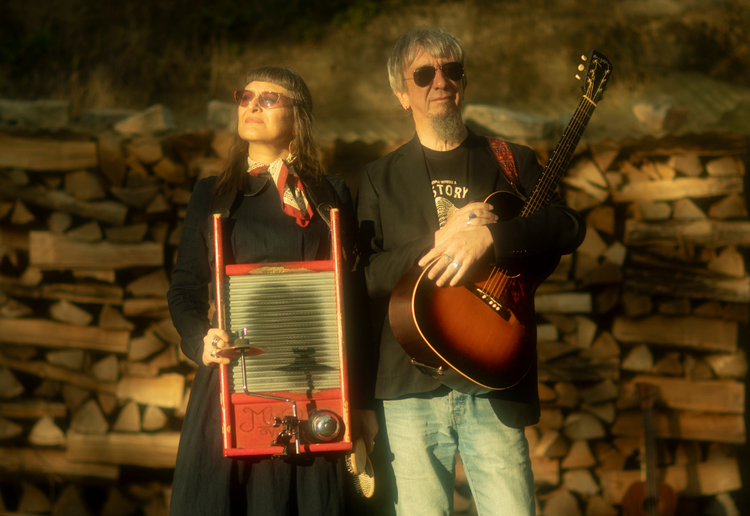 A man and woman stand in front of a countryside depicting background, holding an acoustic guitar and a wash board instrument