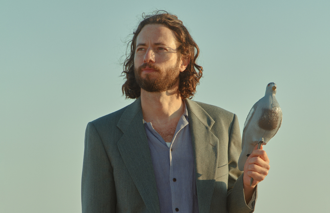 A man in a grey suit holds a model of a pigeon in one hand against a blue sky