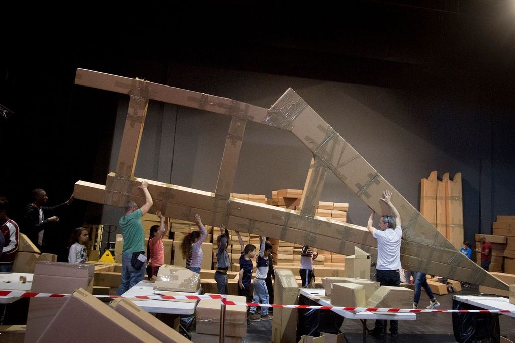 In a dark room, a crowd of people hold up a makeshift bridge piece, constructed from lots of cardboard