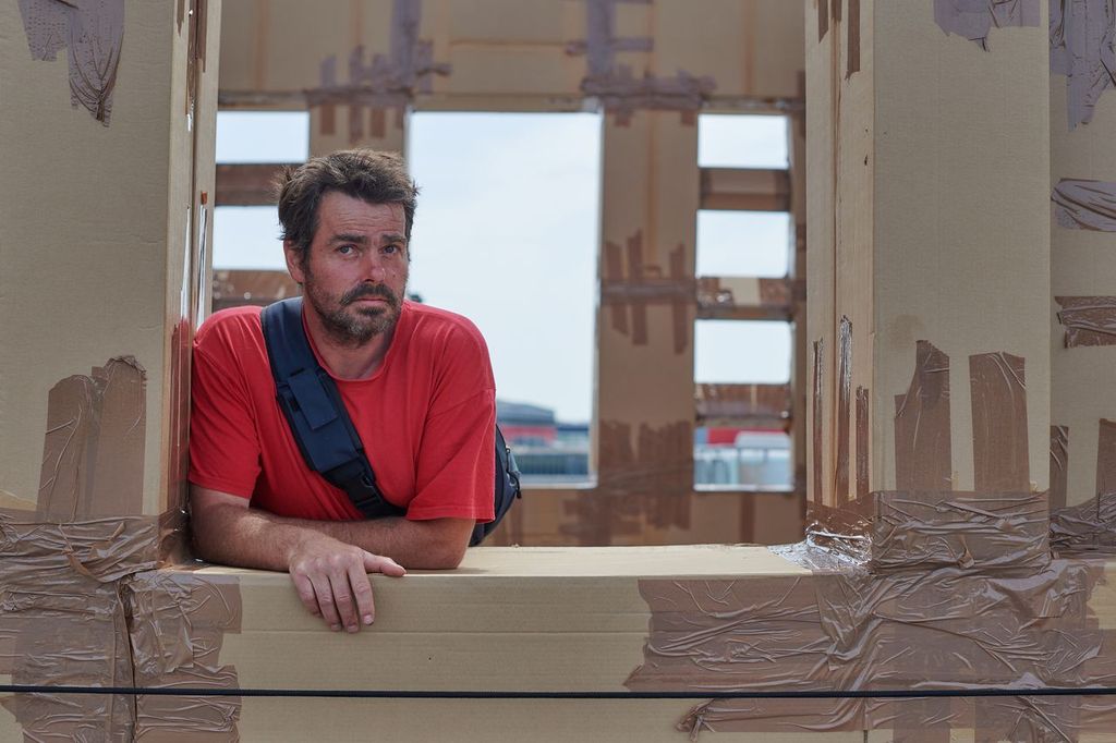 A man in a red shirt stands surrounded by a cardboard structure