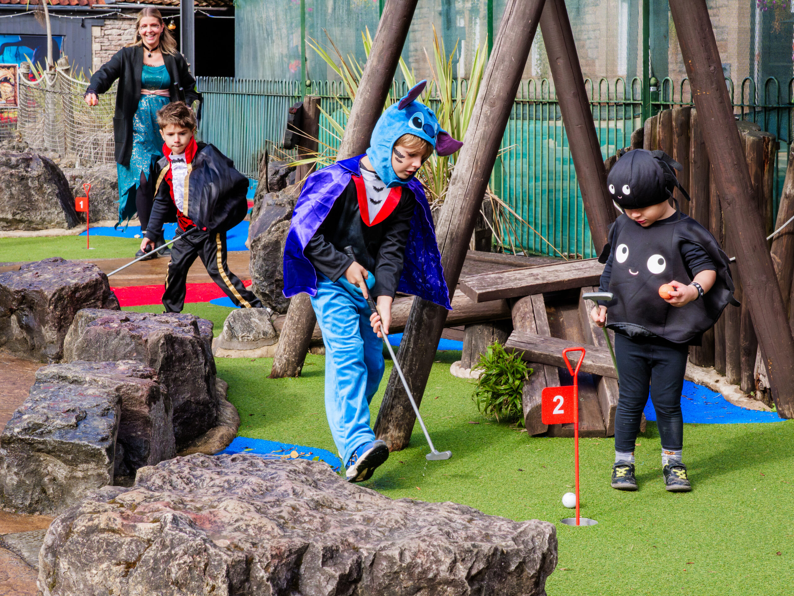 3 children dressed in Halloween costumes (two vampires and a spider) play mini golf