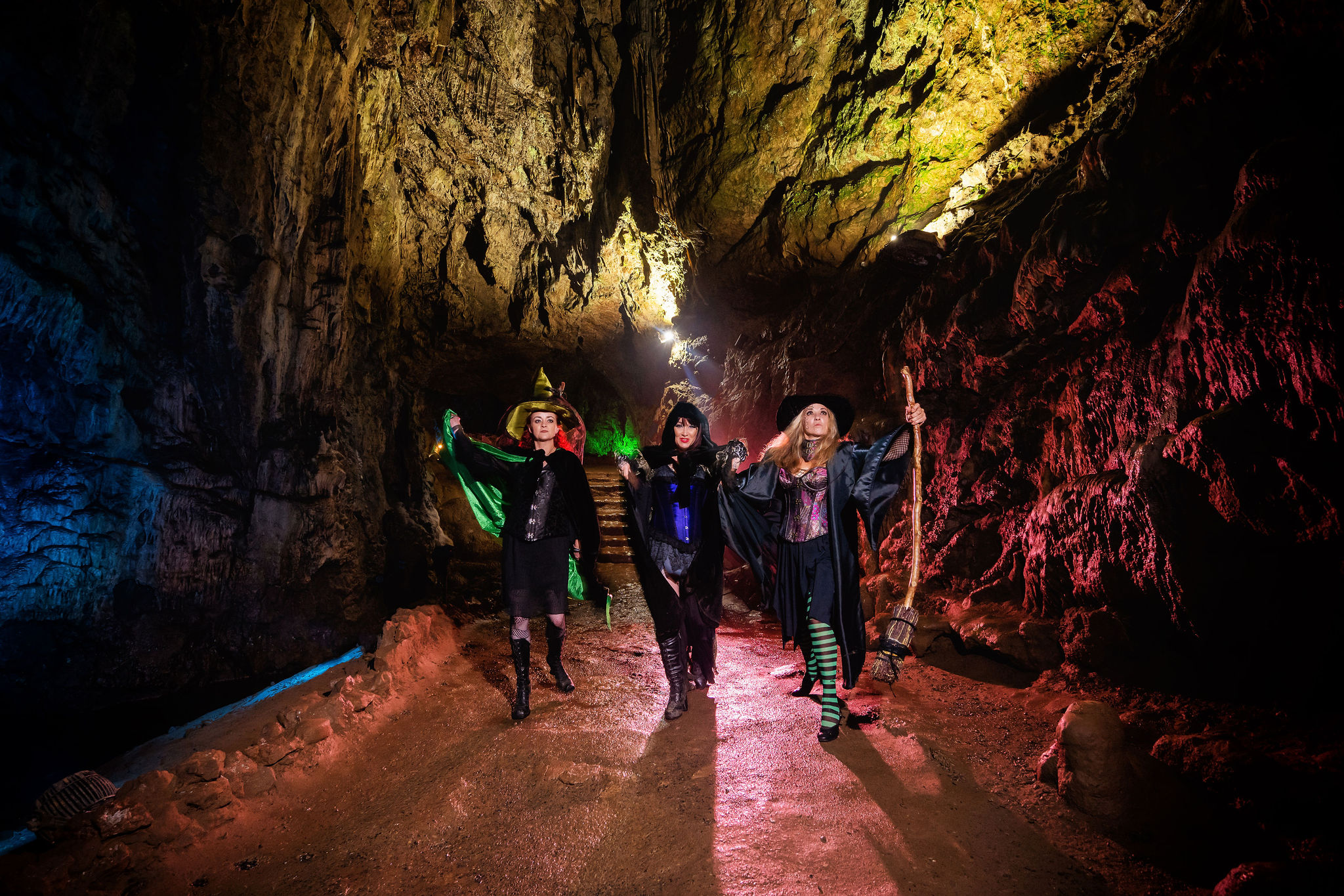 3 women dressed as witches pose in part of Wookey Hole cave, blue pink and yellow lights project onto the cave walls