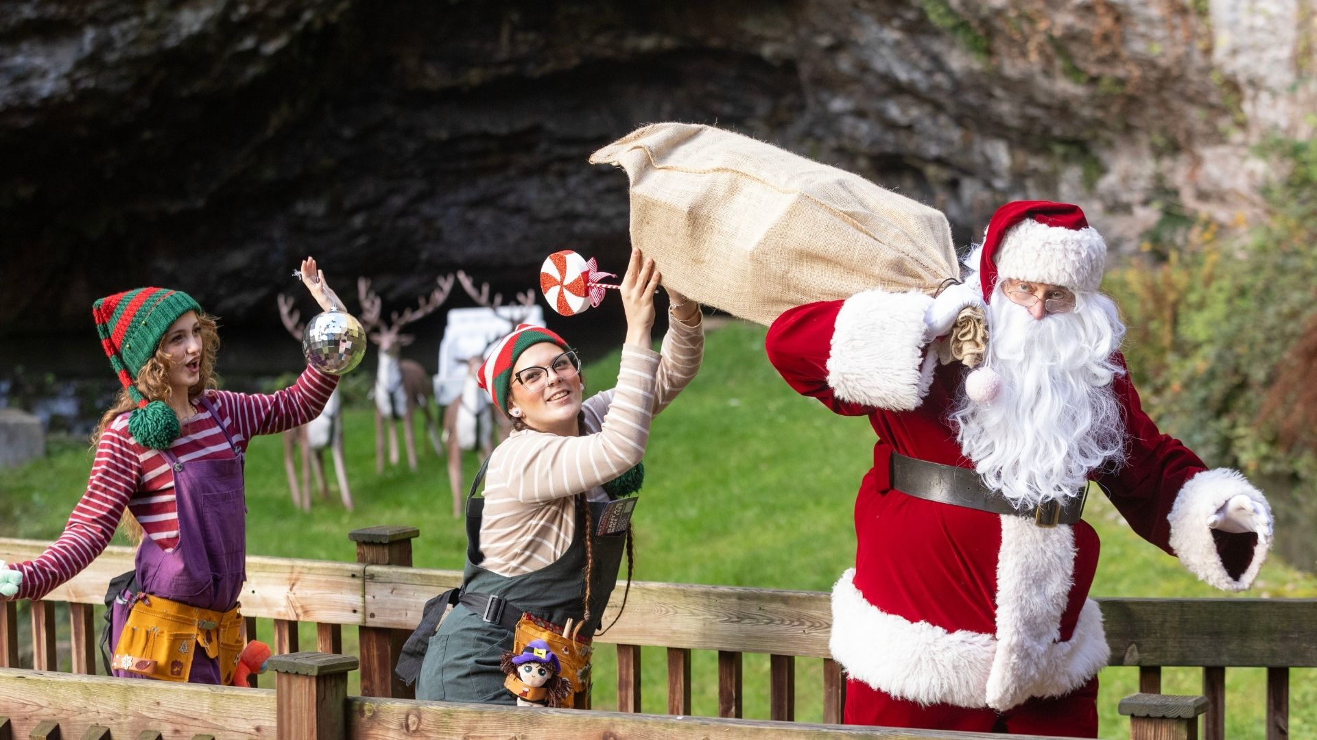Santa walks with a bag over his shoulder and two elves behind him. In the background green grass