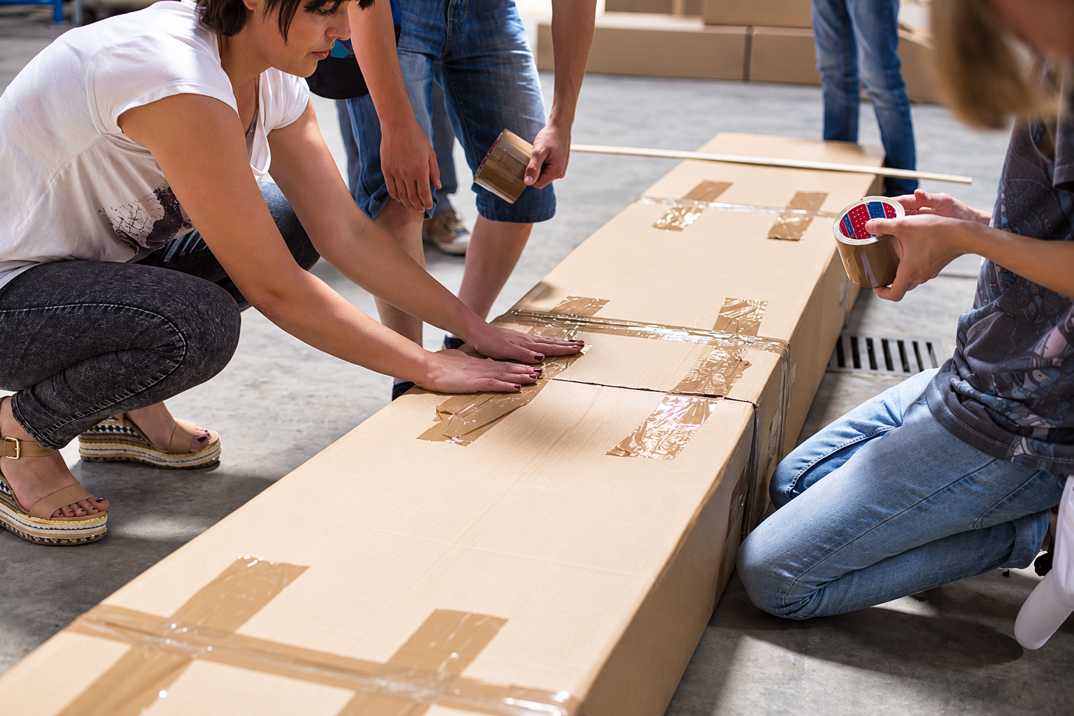 A woman and youngters crouching down to make a cardboard structure with sticking tape.
