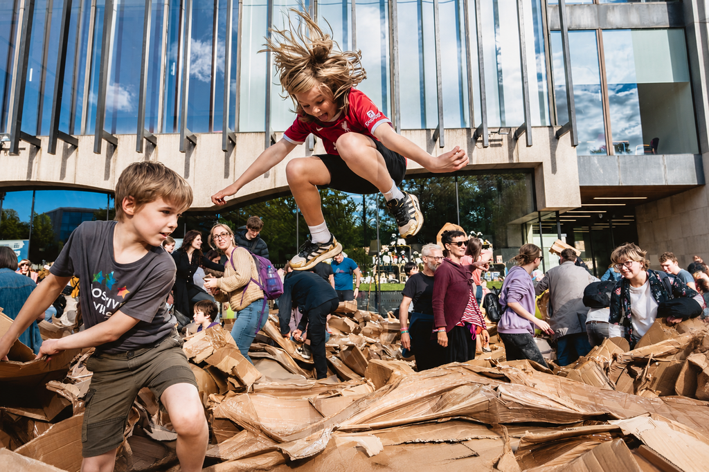 Two Children playing with cardboard, a boy and a girl. The girl is jumping onto of a pile of flat cardboard, stacked on top of each other.