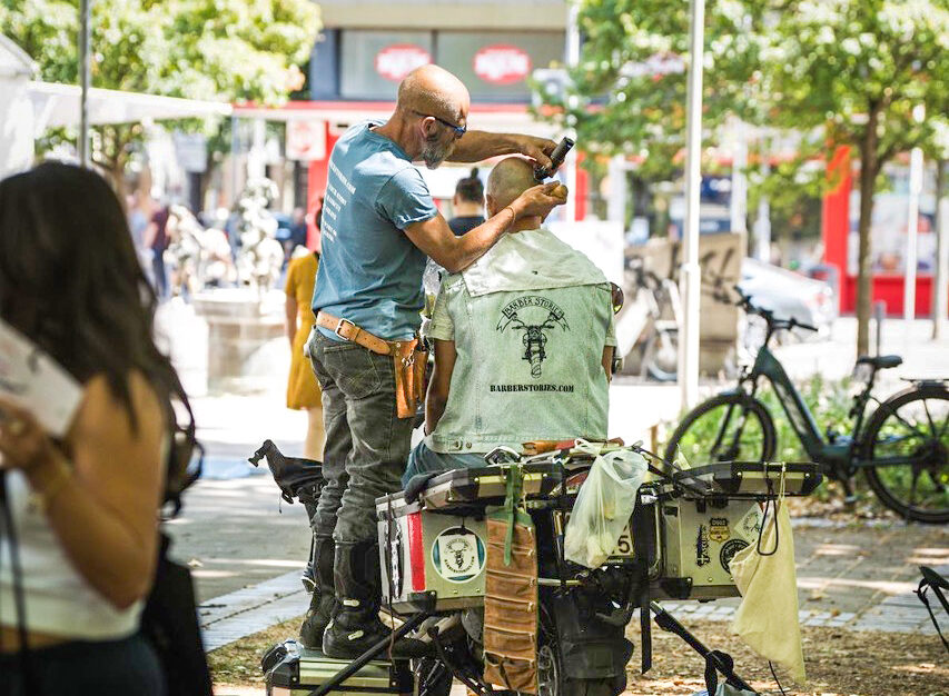 A barber standing on a box giving a haircut to a man sitting on a motorbike