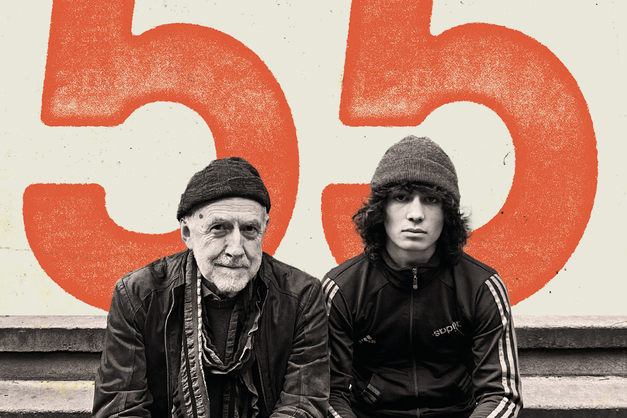 An elderly man and a teenager sit by side to face the camera for a black and white photo in front of a cream and red background