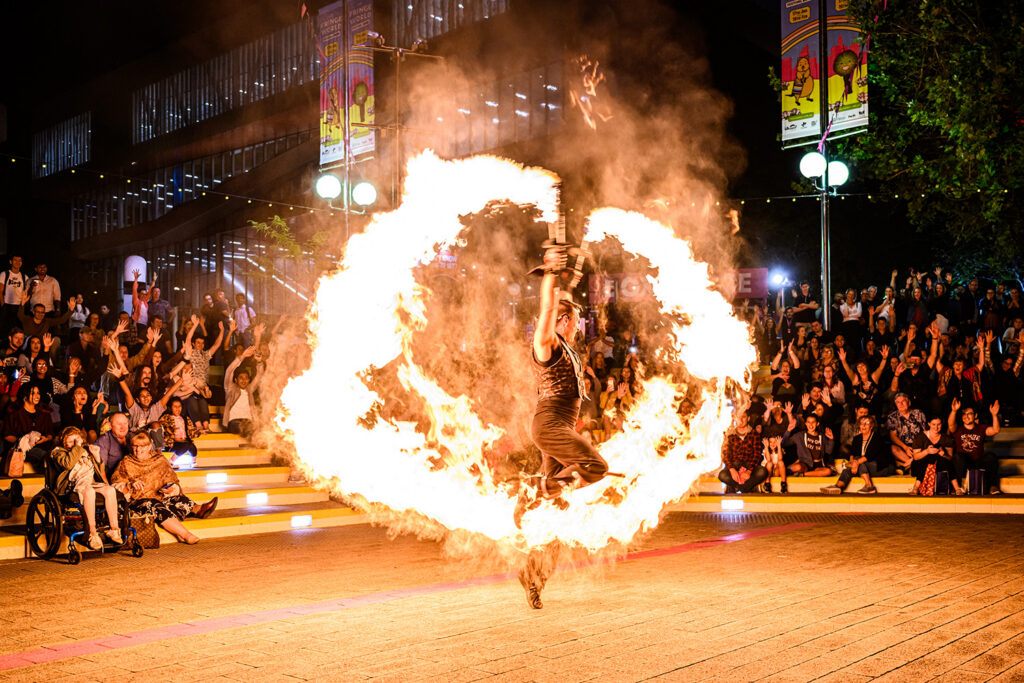 A dancing man holding two flaming torches standing inside a ring of fire while being watched by a large crowd
